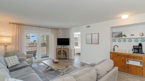 A modern living room with a sofa, coffee table, and a TV. There is a balcony with a view, a kitchen area with wooden cabinets, and a dining table. - Steps to Beach! Spacious 3bd with pool! Murrells Inlet! Family friendly (Murrells Inlet)