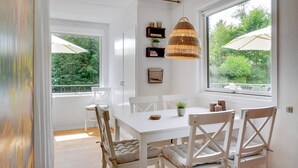A dining area with a white table and chairs, a wicker pendant light, and a view of greenery through large windows. - Cozy cottage near the beautiful Rågeleje beach. (Vejby)