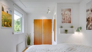 A bedroom with a large bed, a wooden wardrobe, a radiator, a potted plant, and wall-mounted shelves with decorative items. - Cozy cottage near the beautiful Rågeleje beach. (Vejby)