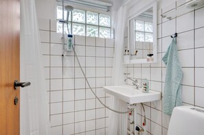 Casa de banho com chuveiro, pia e sanita, apresentando azulejos brancos e uma janela com vista.