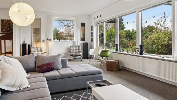 A modern living room with a grey sectional sofa, a white coffee table, and a large window with a view of greenery. - Large summer house in Bandholm on Lolland (Bandholm)