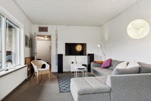A modern living room with a grey sofa, a wicker chair, a small table, and a TV mounted on the wall. - Large summer house in Bandholm on Lolland (Bandholm)