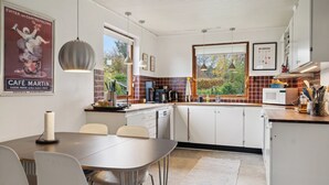A modern kitchen with white cabinets, a dining table with chairs, pendant lights, and a window with a view of trees. - Large summer house in Bandholm on Lolland (Bandholm)