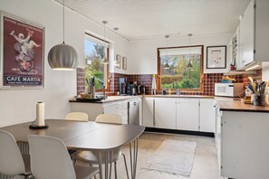 A modern kitchen with white cabinets, a dining table with chairs, pendant lights, and a window with a view of trees.
