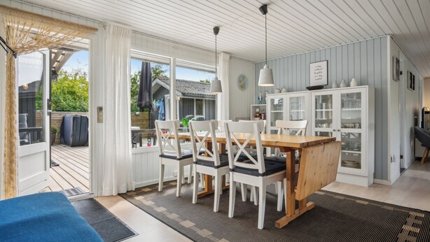 Un comedor con una mesa de madera y sillas blancas, una cocina con armarios blancos, y una vista a través de la puerta de cristal.