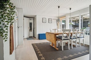 A dining area with a wooden table and white chairs, a patterned rug, and a view through large windows. - Lovely cottage in Pøt Strandby (Juelsminde)