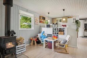 A cozy room with a wood-burning stove, a rocking chair, a dining table, and a window with a view of greenery. - Lovely cottage in Pøt Strandby (Juelsminde)