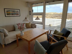 A room with a wooden table, a sofa, and a chair. There is a window with a view of the outdoors. - Lovely summer house on natural grounds (Frøstrup)