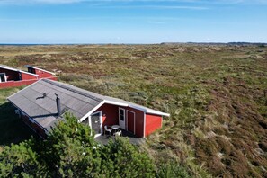 A red building with a flat roof, a small patio with a table and chairs, and a view of a barren landscape. - Lovely summer house on natural grounds (Frøstrup)