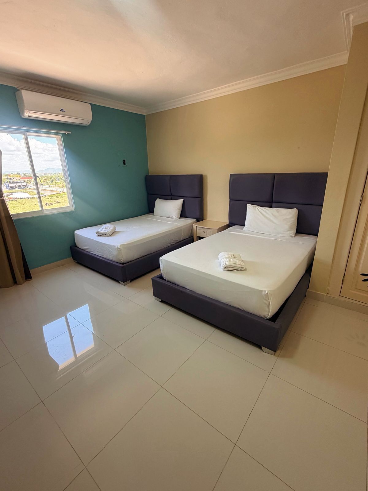 A hotel room with two beds, a window, and an air conditioning unit.