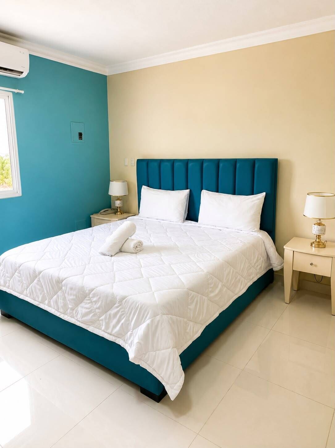 Un dormitorio con cabecera color turquesa, colcha blanca, dos mesitas de noche con lámparas y un equipo de aire acondicionado en la pared.