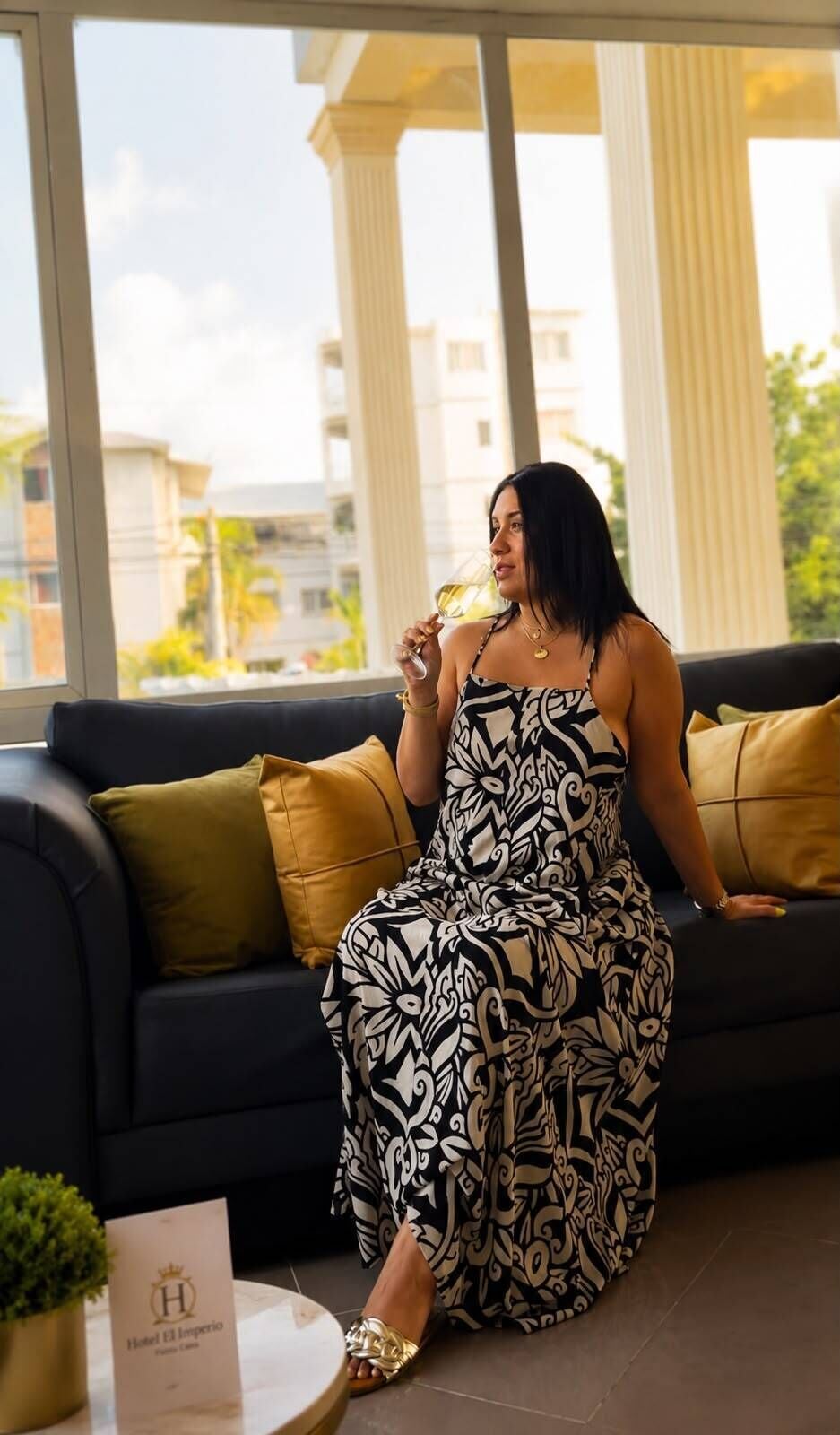 Una mujer sentada en un sofá oscuro con cojines estampados, sosteniendo un vaso.