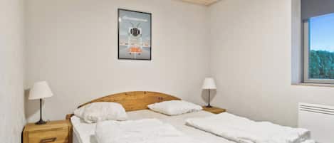 Un dormitorio con cabecera de madera, cama con ropa blanca, dos lámparas de noche, un cartel en la pared y una ventana con vistas a árboles.