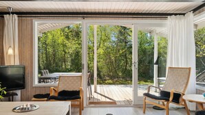 A modern interior with a TV, wooden furniture, and a glass door leading to a deck. - Lovely family cottage near child-friendly beach (Aakirkeby)