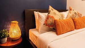 Ein ordentlich bezogenes Bett mit weißen und orangefarbenen Kissen, ein Nachttisch mit Lampe und eine an der Wand montierte Klimaanlage.