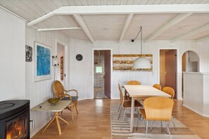A room with a fireplace, dining table, chairs, and a kitchen area visible through an open door. - Lovely summer house with sea view (Haderslev)