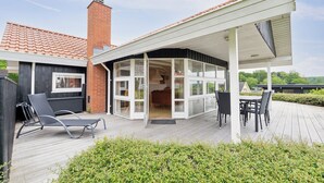 Een overdekt buitengebied met een eettafel en stoelen, een loungestoel en een zicht op het interieur door glazen deuren.