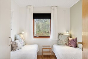 A room with two beds, a window with a view of trees, a small table, and a wall-mounted light.