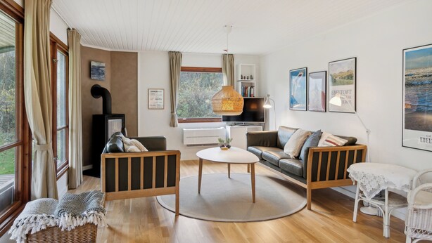 A cozy living room with a sofa, a coffee table, a wicker chair, and a fireplace. There are large windows with curtains, a bookshelf, and framed pictures on the wall. - Cozy cottage steps from Lyngsbæk beach (Ebeltoft)