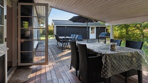 Eine Terrasse im Freien mit Tisch und Stühlen, einem überdachten Bereich und Blick auf ein Gebäude im Hintergrund.