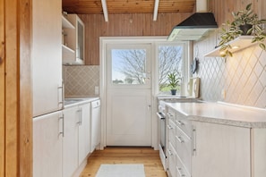 Une cuisine avec des meubles en bois, un plan de travail blanc, une cuisinière et une fenêtre donnant sur des arbres.