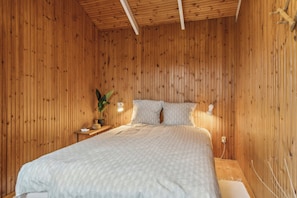 A wooden-paneled room with a bed, a nightstand, a lamp, and a potted plant.