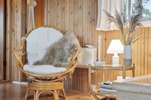 Una silla de mimbre con un cojín mullido, una mesa con una lámpara y un florero con plantas secas, y paneles de madera en las paredes.