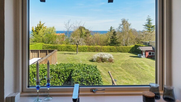 A window with a view of a well-maintained garden, a wooden deck, and a house in the background. - Modern holiday home with sea view in Tejn (Allinge-Sandvig)