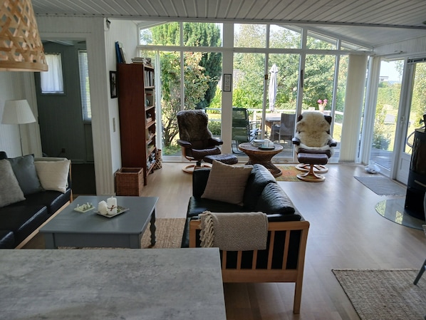 A spacious living room with a large window, a sofa, a chair, a bookshelf, and a dining area.