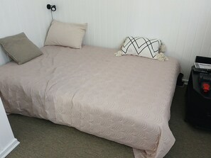 Ein Bett mit einer beigen Steude, zwei Kissen, einem schwarz-weißen Kissen mit Muster und einem kleinen schwarzen Tisch mit Fernbedienung.