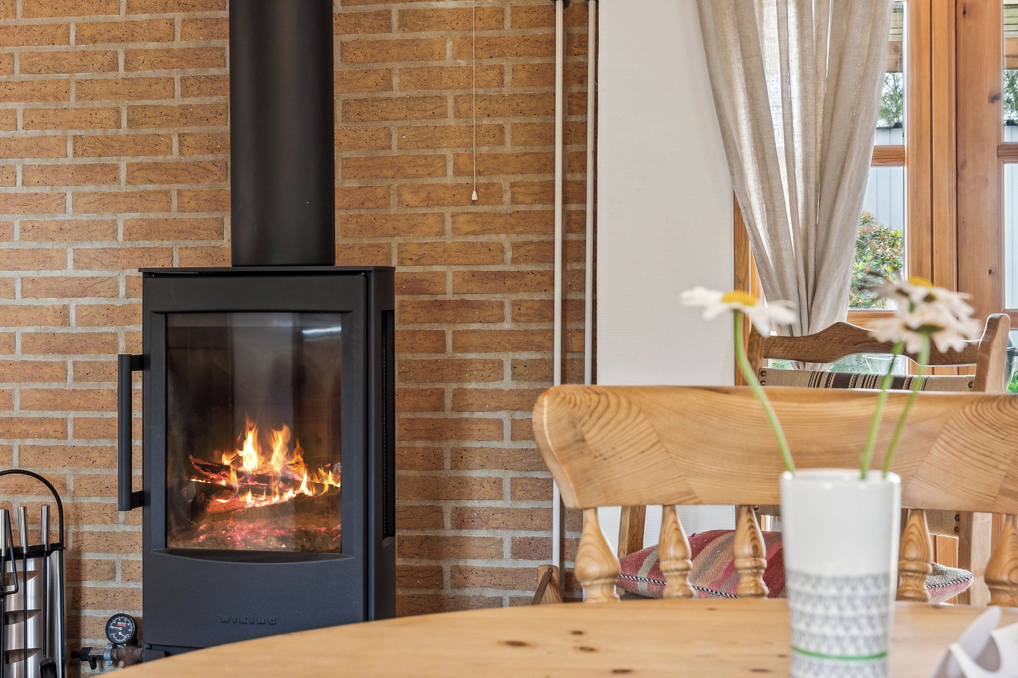 A wood-burning stove, a brick wall, a wooden table, and a chair with a vase of flowers.