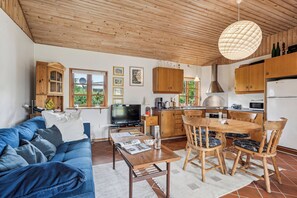 Een gezellige woonkamer met een blauwe bank, een houten eettafel en stoelen, een keuken met houten kasten, en een groot raam.