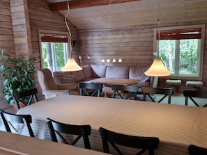 Et hyggeligt sommerhusinteriør med et spisebord, stole, en sofa og to vinduer med persienner.