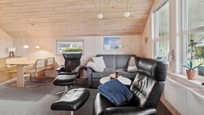 Ett mysigt rum med ett träpanelat tak, en grå soffa, en svart läderfåtölj, ett träbord med stolar och ett fönster med utsikt över grönområden.
