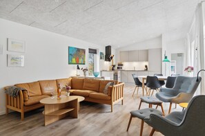 A modern living room with a sofa, coffee table, and chairs, a kitchen in the background, and artwork on the walls.