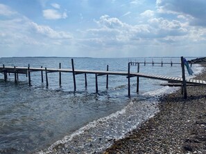 A wooden pier extending into calm waters, surrounded by pebbles and a clear sky.