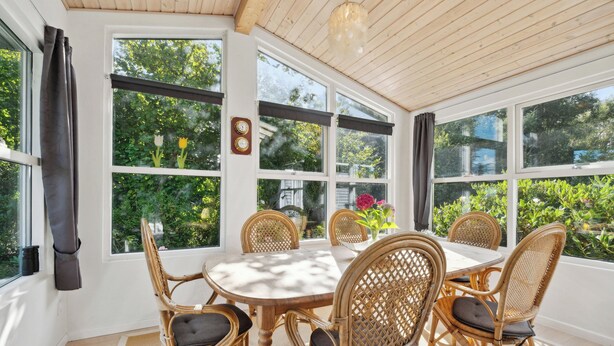 Une pièce lumineuse, avec une grande table et des chaises en osier, de grandes fenêtres et un ventilateur de plafond.