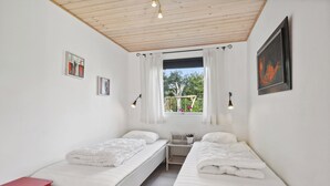 Une petite chambre propre, avec deux lits simples, une chaise rouge, une petite table et une fenêtre avec des rideaux.