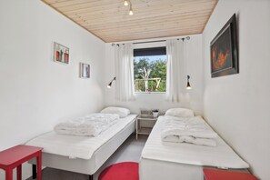 Una habitación pequeña y limpia con dos camas individuales, una silla roja, una mesita y una ventana con cortinas.