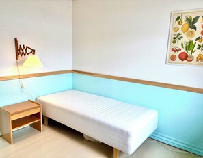 A small, single-bed room with a wooden nightstand, a wall-mounted lamp, and a fruit illustration on the wall.