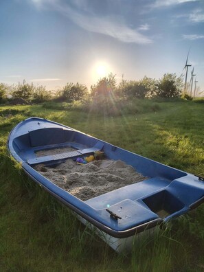 A small blue boat with a flat bottom, resting on grass with sand inside, and wind turbines in the background.