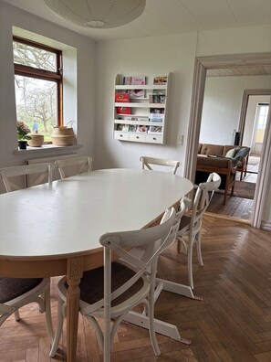 Un comedor con una mesa y sillas blancas, una estantería con libros y objetos decorativos, y un ventanal con vistas a árboles.