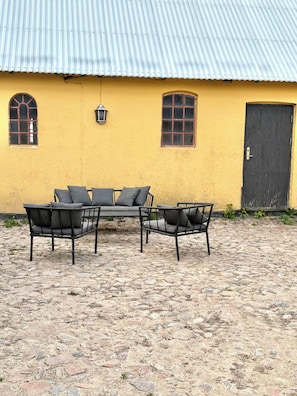 Un edificio amarillo con techo de metal corrugado, dos ventanas arqueadas, una puerta y un patio empedrado con una mesa y sillas de metal.