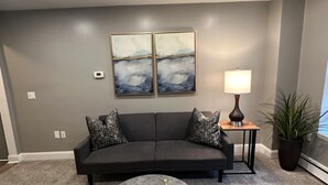 Una sala de estar moderna con un sofá gris, dos cuadros abstractos, una mesa de centro redonda, una lámpara de pie y una planta en maceta.