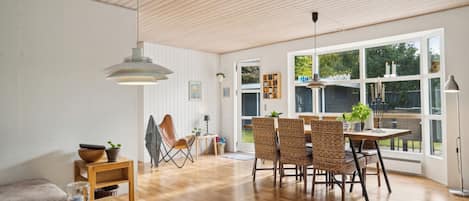 A spacious room with a wooden ceiling, large windows, a dining area with wicker chairs, and a table with fruit.