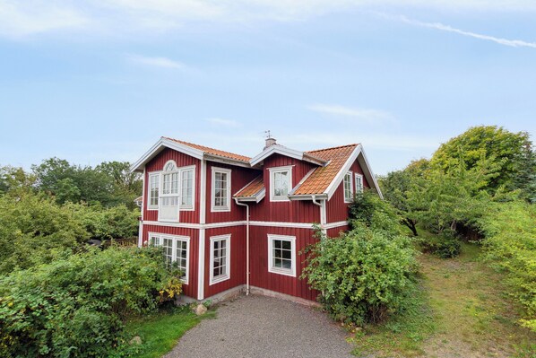 Une maison rouge à deux étages, avec une allée en gravier et entourée de verdure.