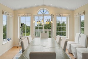 A spacious dining room with a long table, white chairs, and large windows with curtains.