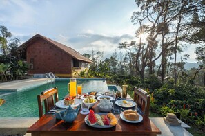 A breakfast table set with various dishes and drinks by a poolside, with a house and lush greenery in the background. - Amã Stays & Trails Alpenhof , Coorg (Madikeri)