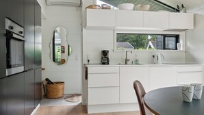 A modern kitchen with white cabinetry, a built-in oven, a coffee maker, and a window above the sink. - Scenic summer house in Asserbo (Frederiksværk)