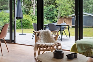 A room with a large glass door leading to an outdoor patio, wicker furniture, a round table with a teapot and cups, and a view of a backyard with greenery and a wooden structure.